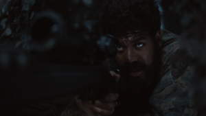 John Stewart With Grown Hair And A Full Beard Looking Through A Rifle Scope.