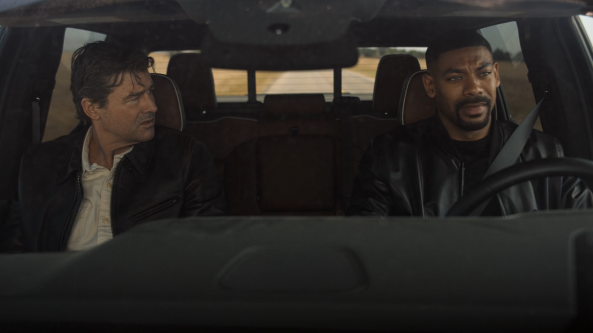 John Stewart Driving A Car With The Halogram Sitting Next To Him.