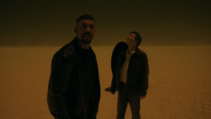 John And The Halogram In A Yellow Desert With A Black Portal Behind Them.